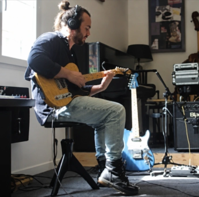 Christopher Chédeville assis jouant de la guitare électrique dans son studio de musique à Linars, casque aux oreilles